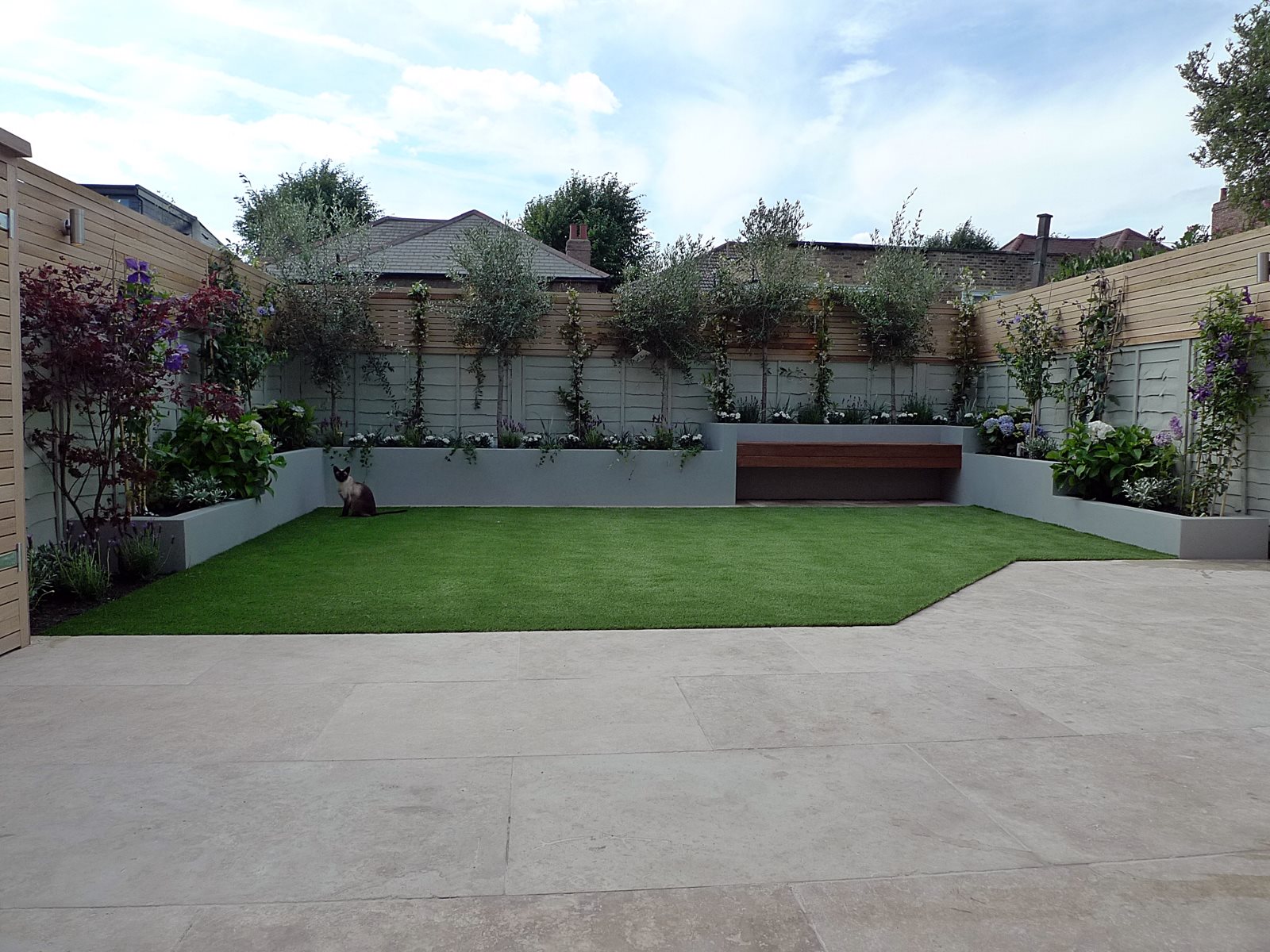 Back garden with light paving stones, artificial grass and a border of decorative plants