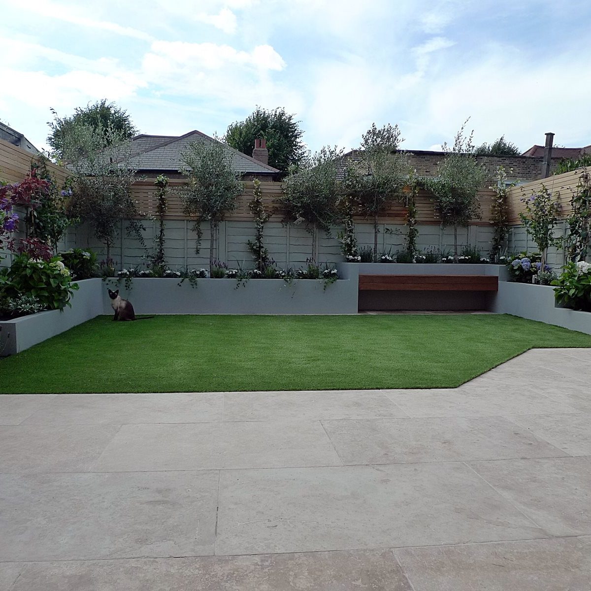 Back garden with light paving stones, artificial grass and a border of decorative plants